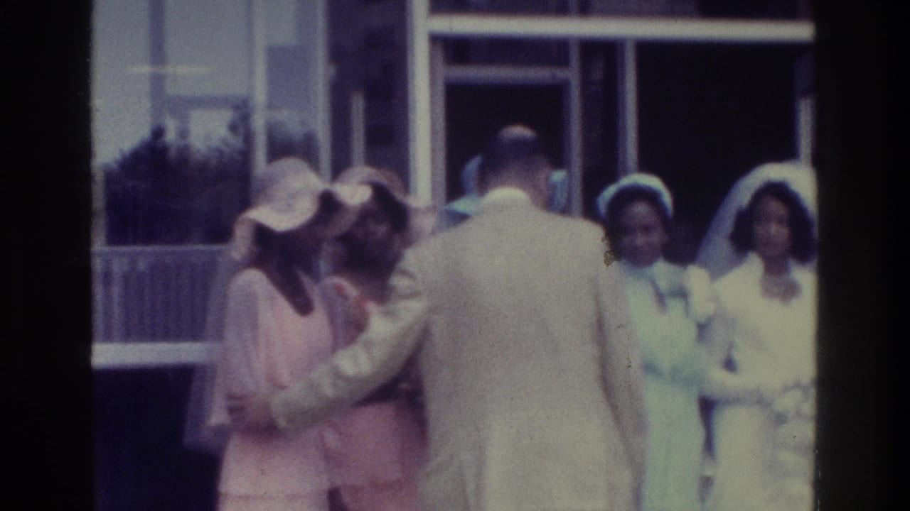 1975 Bronx home-movie frame showing a man positioning bridesmaids in pastel dresses and wide hats near a building doorway.