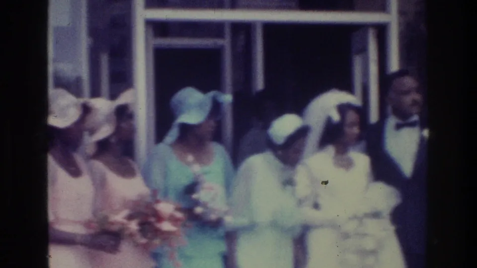 1975 Bronx home-movie frame showing bridesmaids in pastel hats beside the bride in white veil during an outdoor wedding-party grouping.