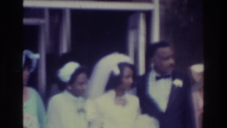 1975 Bronx wedding frame with bride in white veil and groom in dark tuxedo standing near pastel-dressed attendants outside a building.