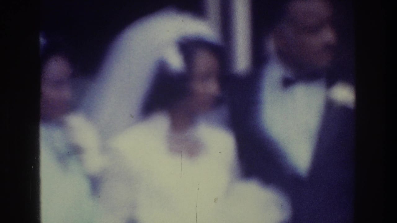 Soft-focus 1975 Bronx home-movie frame showing a bride in a veil beside a groom in a tuxedo indoors.