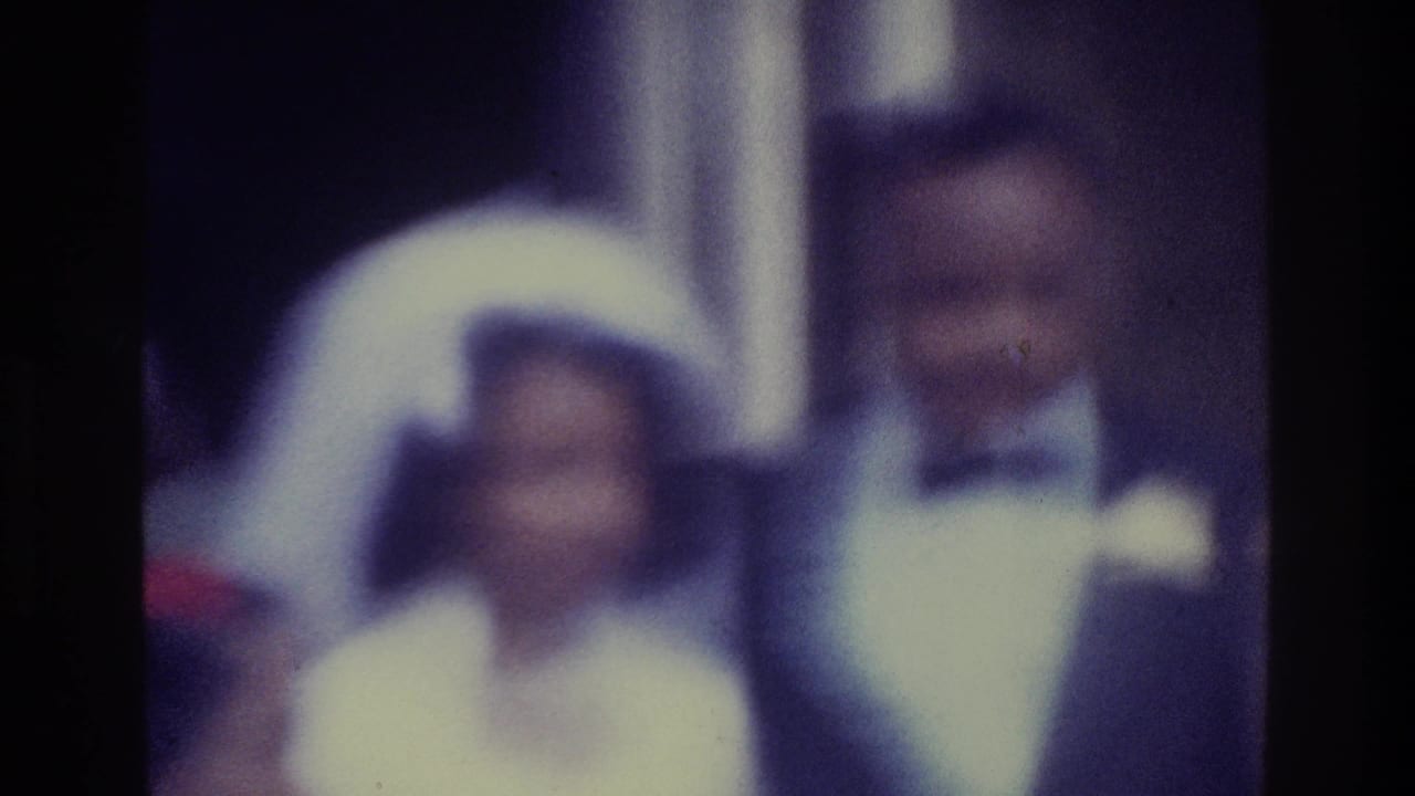 Soft 1975 Bronx 8mm frame of a bride in a white veil standing near a groom in a dark tuxedo at a wedding gathering.