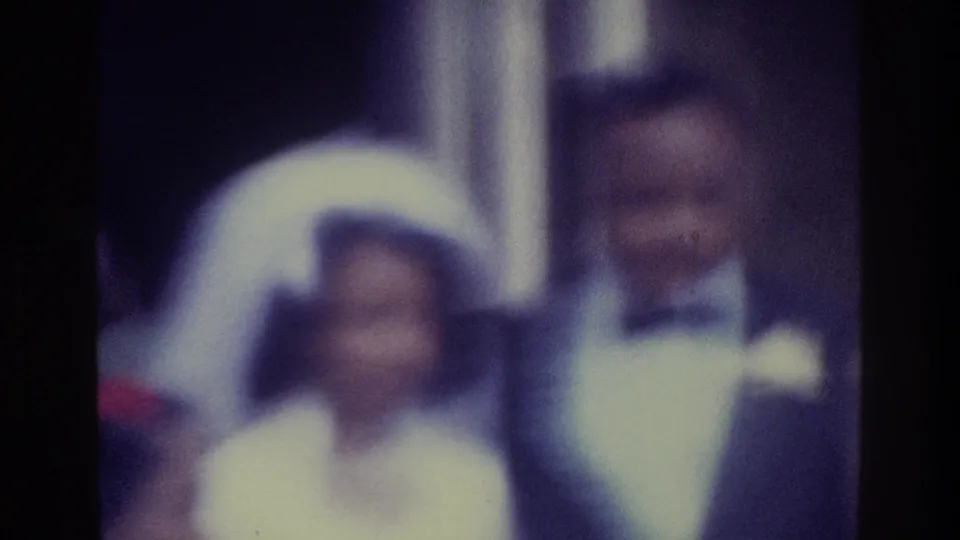 Soft 1975 Bronx 8mm frame of a bride in a white veil standing near a groom in a dark tuxedo at a wedding gathering.