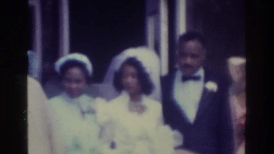 1975 Bronx home-movie frame showing a Black bride in a white veil beside the groom in a tuxedo, with guests nearby.