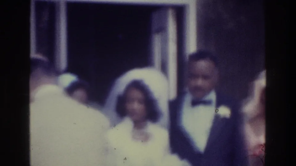 Black wedding couple in 1975 Bronx footage, partly obscured by a guest passing in the foreground near a doorway.