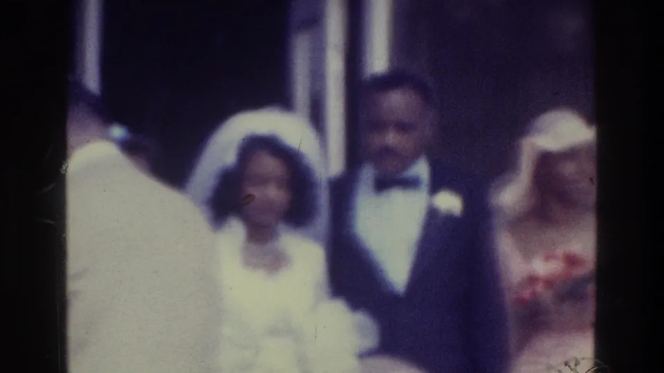 1975 Bronx wedding home movie frame with the bride and groom surrounded by guests, including a woman holding red flowers.