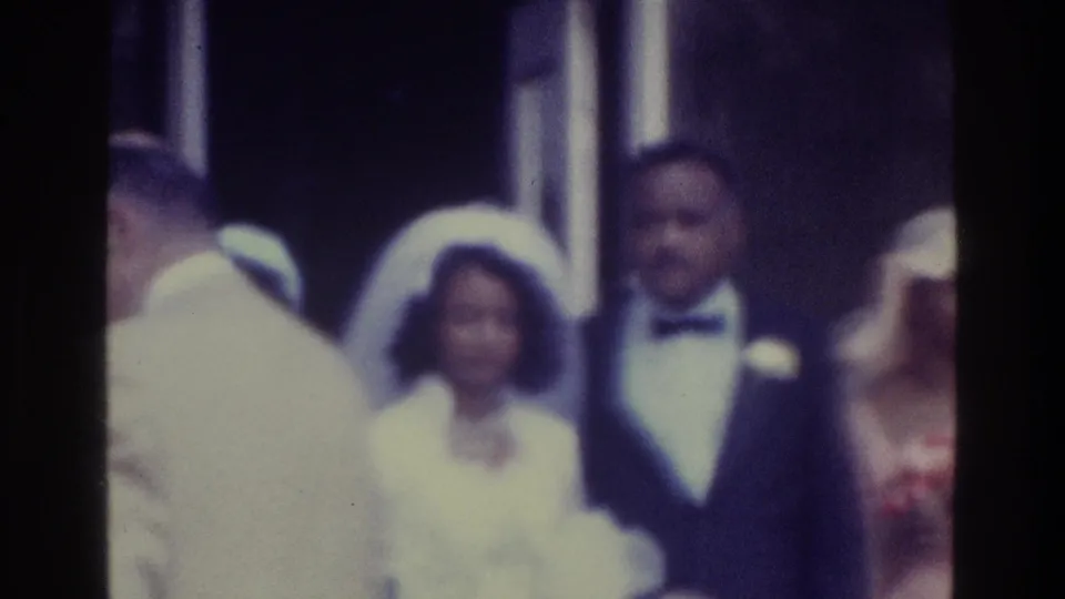 Black bride and groom in 1975 Bronx 8mm footage, standing with wedding guests while a foreground figure blocks part of the scene.