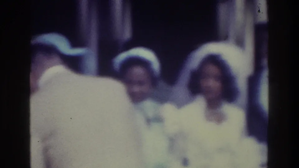 Soft 1975 Bronx 8mm frame showing a groom near formally dressed women, with faces and clothing blurred by focus and exposure.