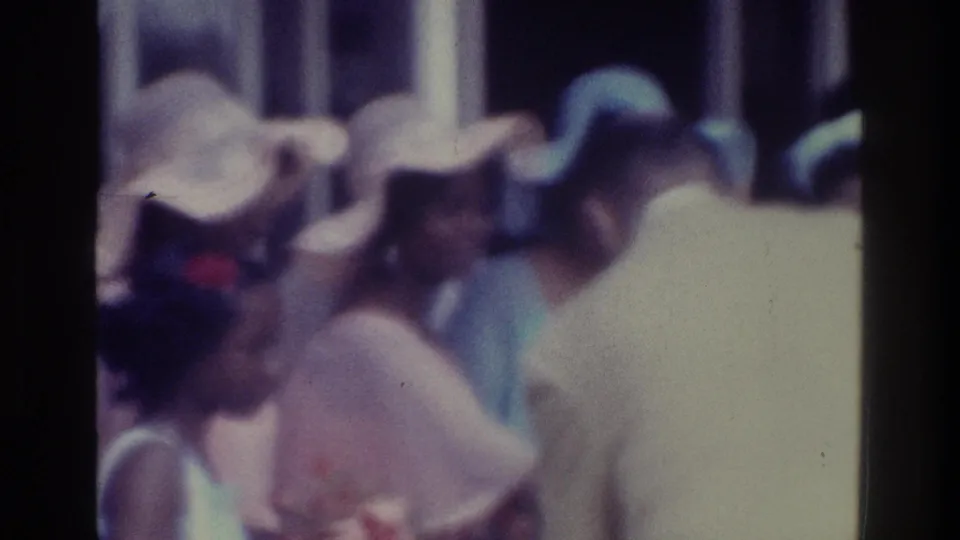 1975 Bronx wedding guests cluster around a veiled bride, with wide-brimmed hats and pale formal clothing visible in soft 8mm color.