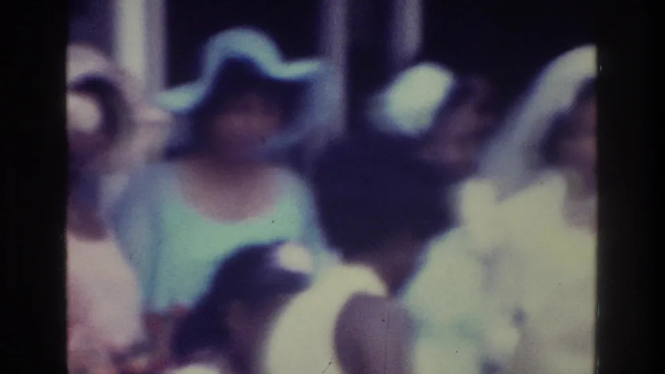 1975 Bronx home-movie frame showing a woman in a pale dress and wide hat among wedding guests, with the groom partly visible nearby.