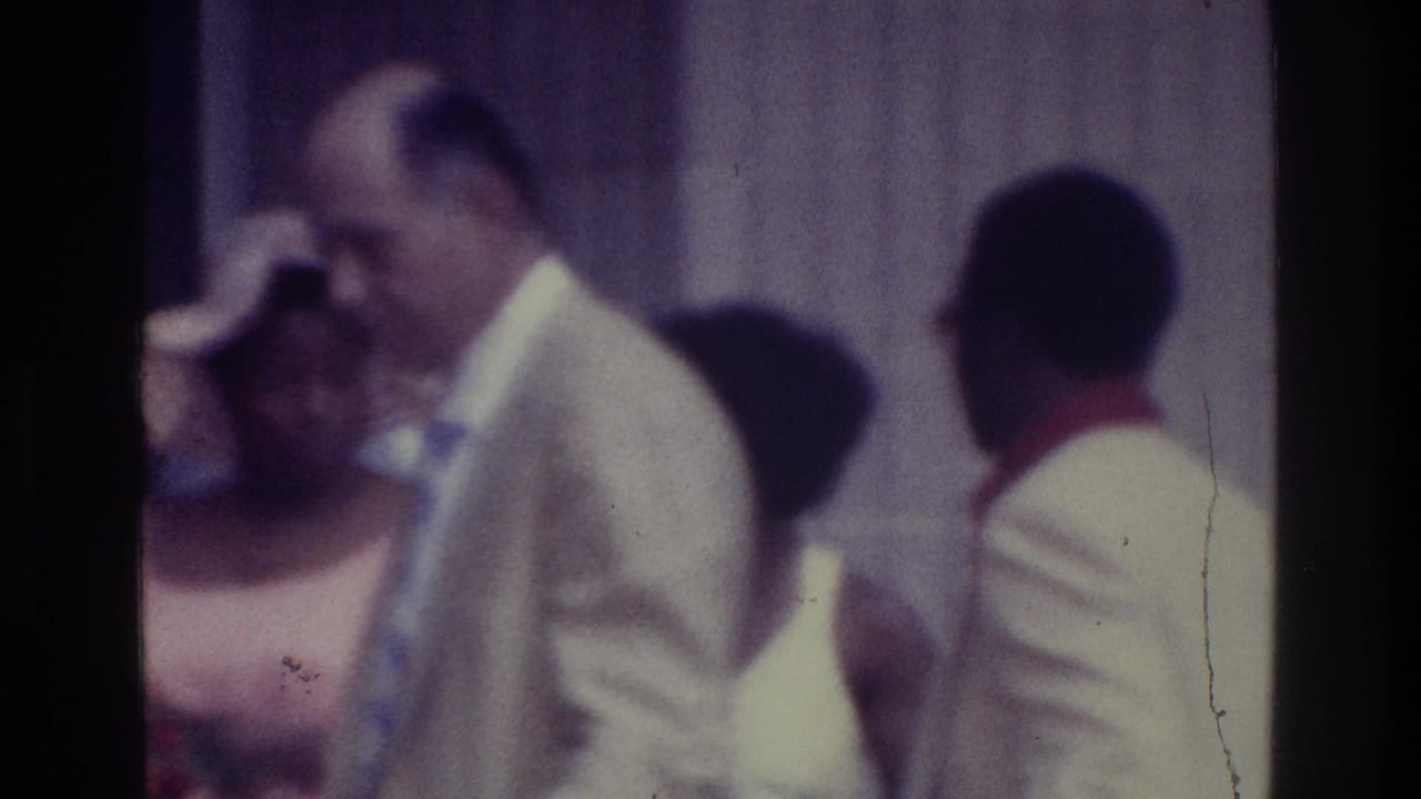1975 Bronx 8mm frame showing adults in suits and dresses gathered near a wall or doorway, with faces soft from motion and focus.