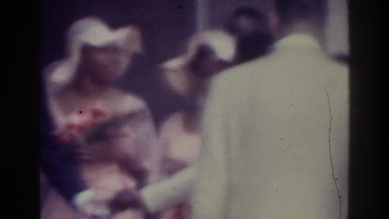 Formally dressed adults face one another, including a pale-hatted woman and a man in a light jacket.