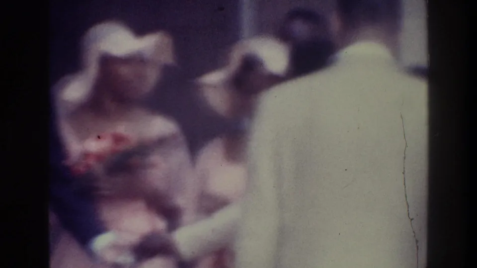 1975 Bronx home-movie frame with adults in formal attire, a pale-hatted woman near center, and a man in a light jacket in the foreground.