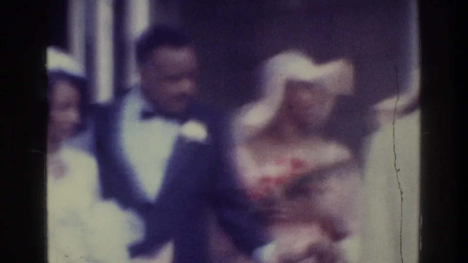 1975 Bronx 8mm frame showing a man in a dark suit and bow tie beside women in pale formal clothing, with soft focus and handheld framing.