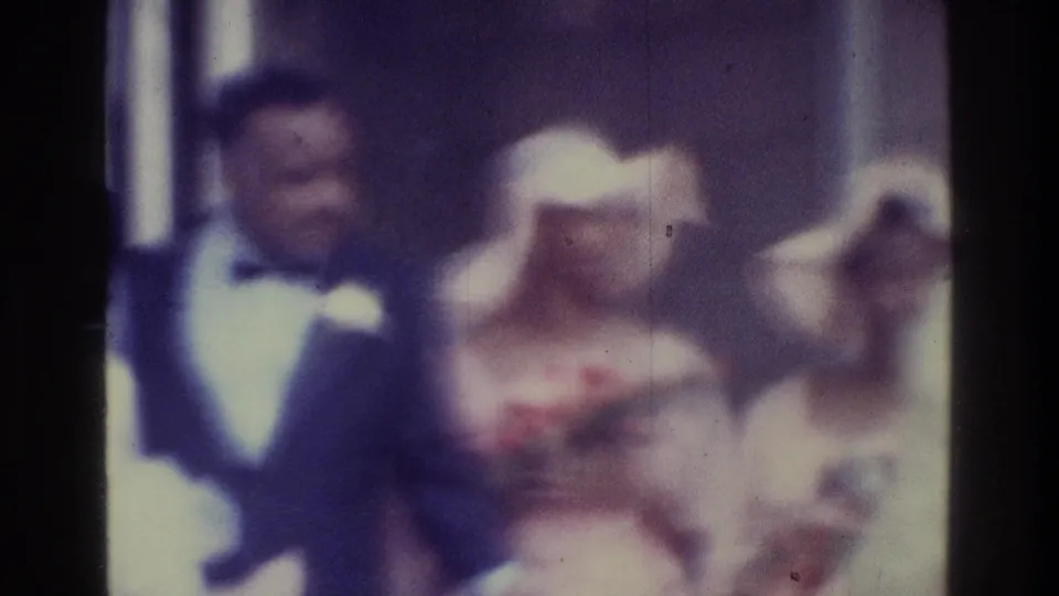 1975 Bronx home-movie frame with a bow-tied man at left, a pale-hatted woman near center, and other adults partly visible at the edge.