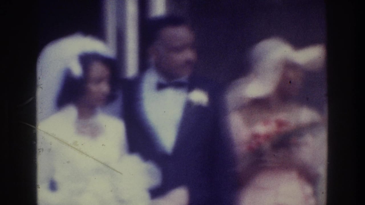 In supported 1975 Bronx home-movie footage, a bride in a white gown and veil stands beside a groom in a dark tuxedo as guests gather close around them.