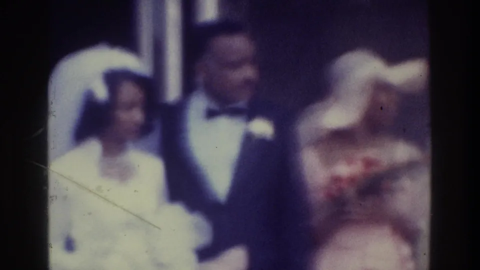 In supported 1975 Bronx home-movie footage, a bride in a white gown and veil stands beside a groom in a dark tuxedo as guests gather close around them.
