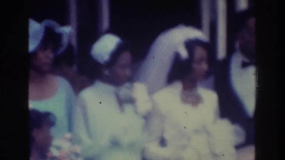 Guests in pale dresses and hats stand near the veiled bride, with the groom partially visible at the edge of the crowded 1975 Bronx wedding frame.