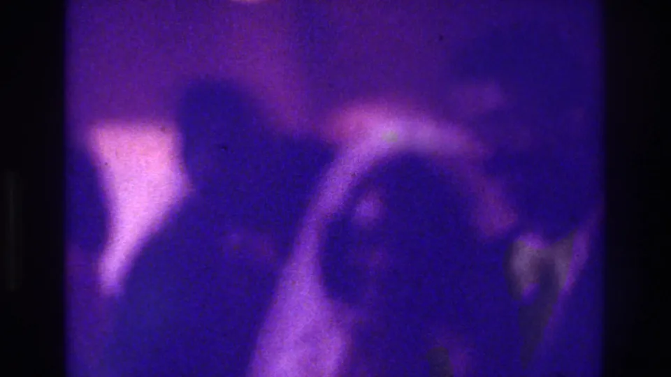 1975 Bronx wedding home-movie frame showing blurred figures near the aisle, with visible grain and strong purple-blue color shift.