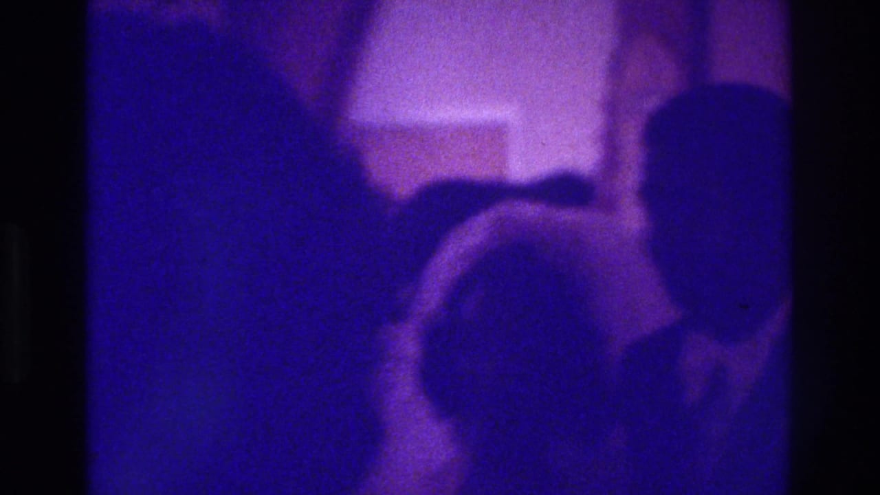 1975 Bronx home-movie frame showing purple-tinted silhouettes of guests gathered indoors at a likely wedding or celebration.