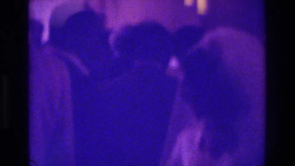 1975 Bronx home-movie frame showing a crowded, purple-tinted interior with guests seated closely at a likely reception.
