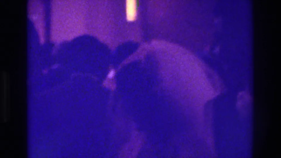 1975 Bronx frame showing a light-haired guest in the foreground and other guests in a dim purple-tinted private interior.