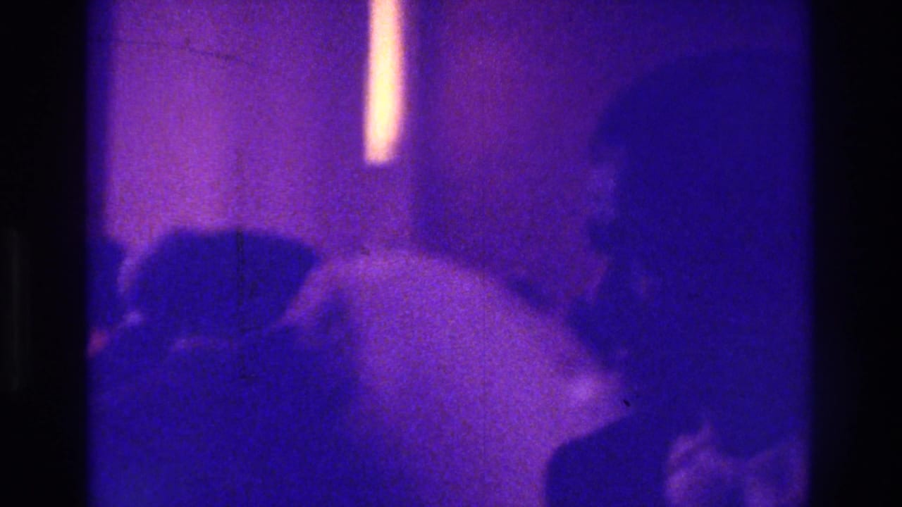 1975 Bronx home-movie frame showing a bride in a white dress among silhouetted wedding guests in a dim interior.