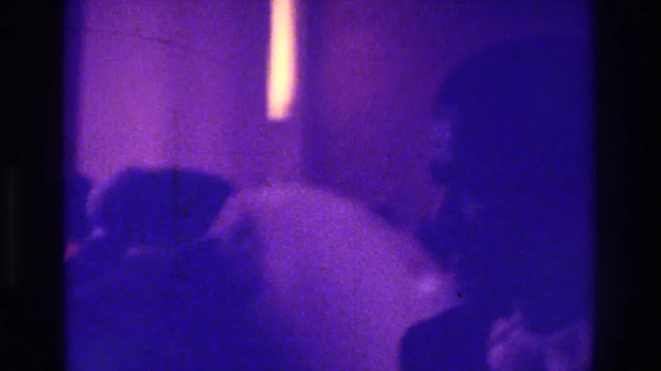 1975 Bronx home-movie frame showing a bride in a white dress among silhouetted wedding guests in a dim interior.