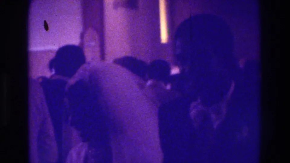 1975 Bronx wedding home-movie frame with the bride turned away, surrounded by seated family members in low light.