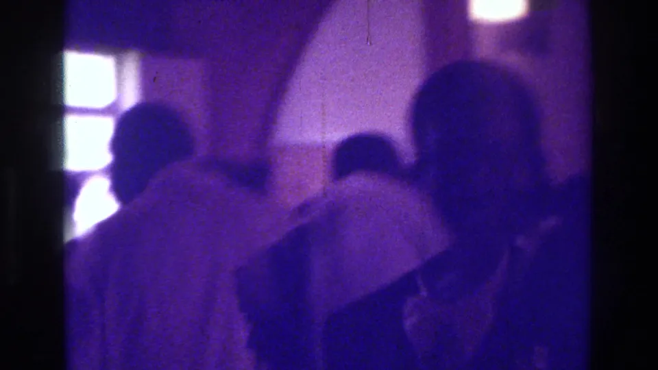 1975 Bronx home-movie frame showing the bride and several guests near bright window or doorway panels in a dim wedding room.