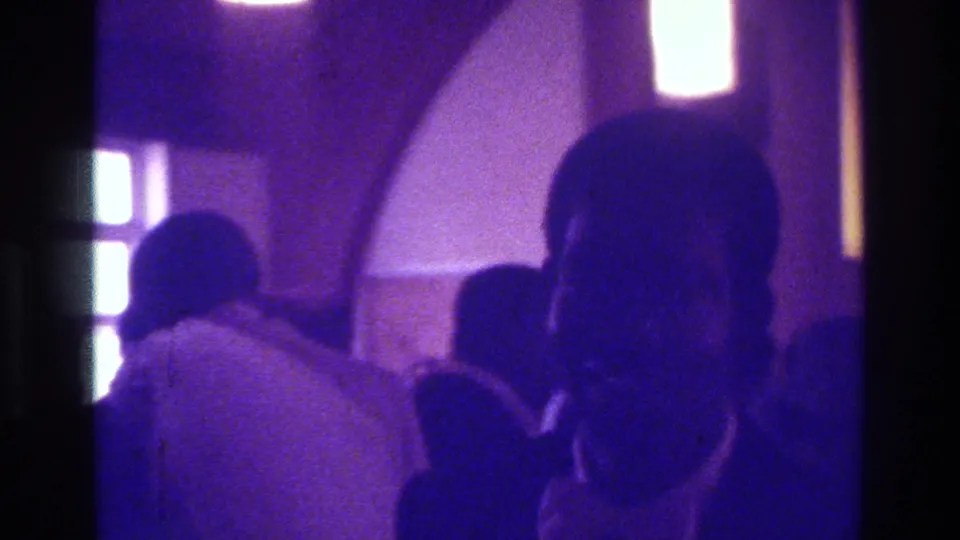 1975 Bronx wedding home-movie frame with the groom in a dark suit at right and the bride and guests behind him.