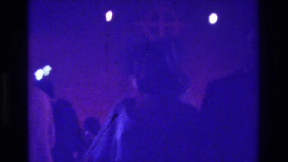 Low-light 1975 Bronx church footage shows indistinct congregants and small bright lights within a purple-tinted interior.
