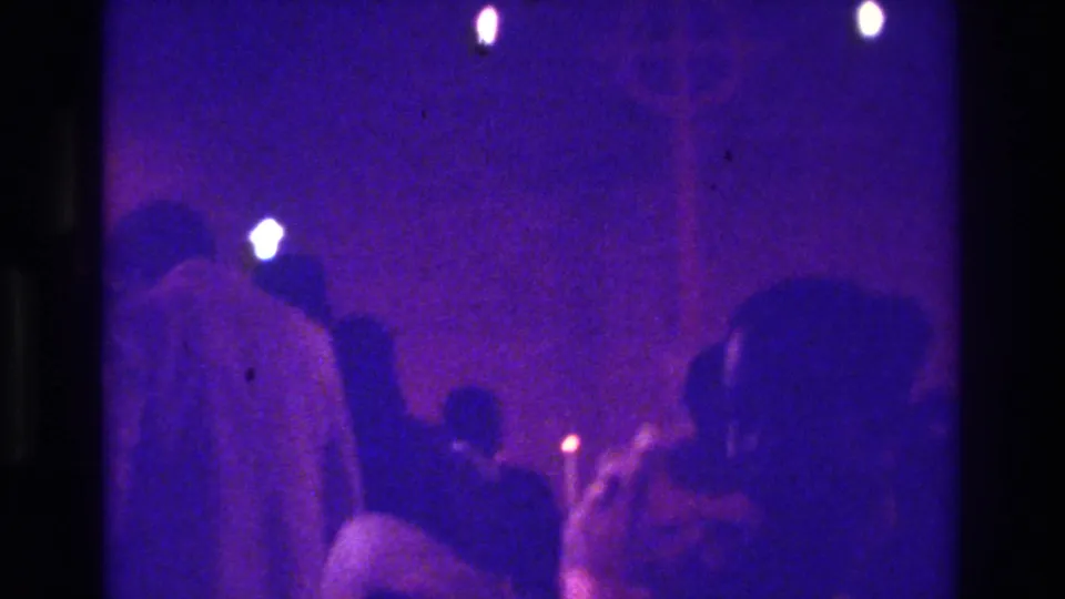1975 Bronx 8mm frame showing worshippers in a dark church interior lit by small candle flames and purple-blue exposure.