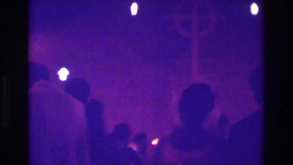 1975 Bronx candlelit church-service frame showing seated silhouettes and small points of flame in a grainy blue-purple image.