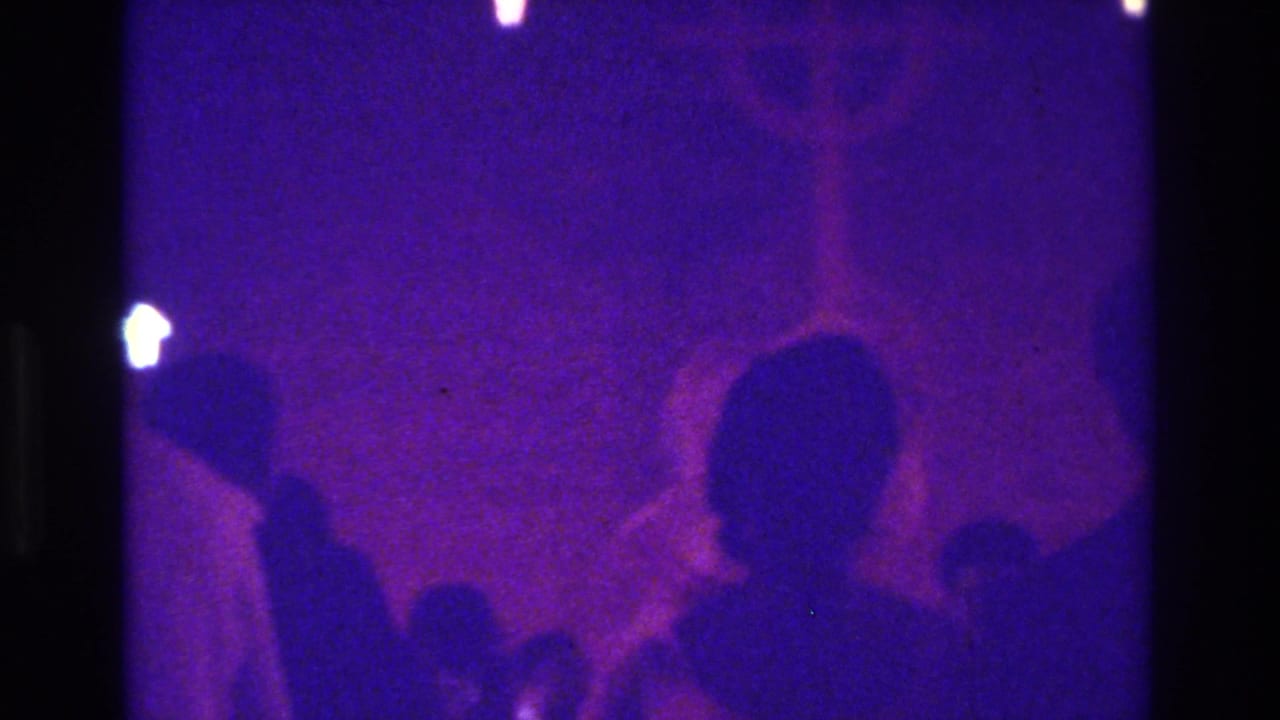 1975 Bronx 8mm frame with purple lighting, dark attendee silhouettes, and a faint bright shape near the top edge.