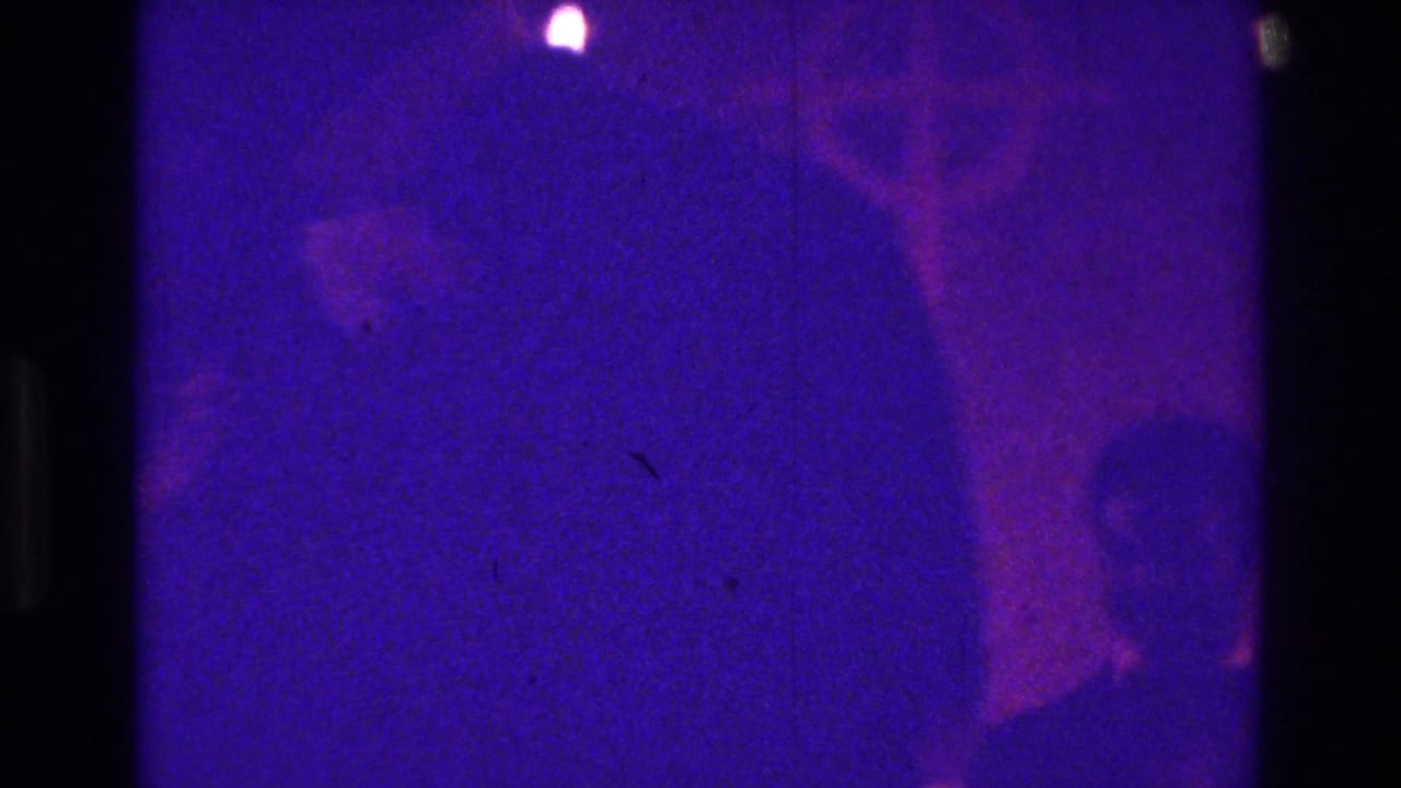 Purple-lit interior with a large cross shape and dark silhouetted figures near the frame edge.