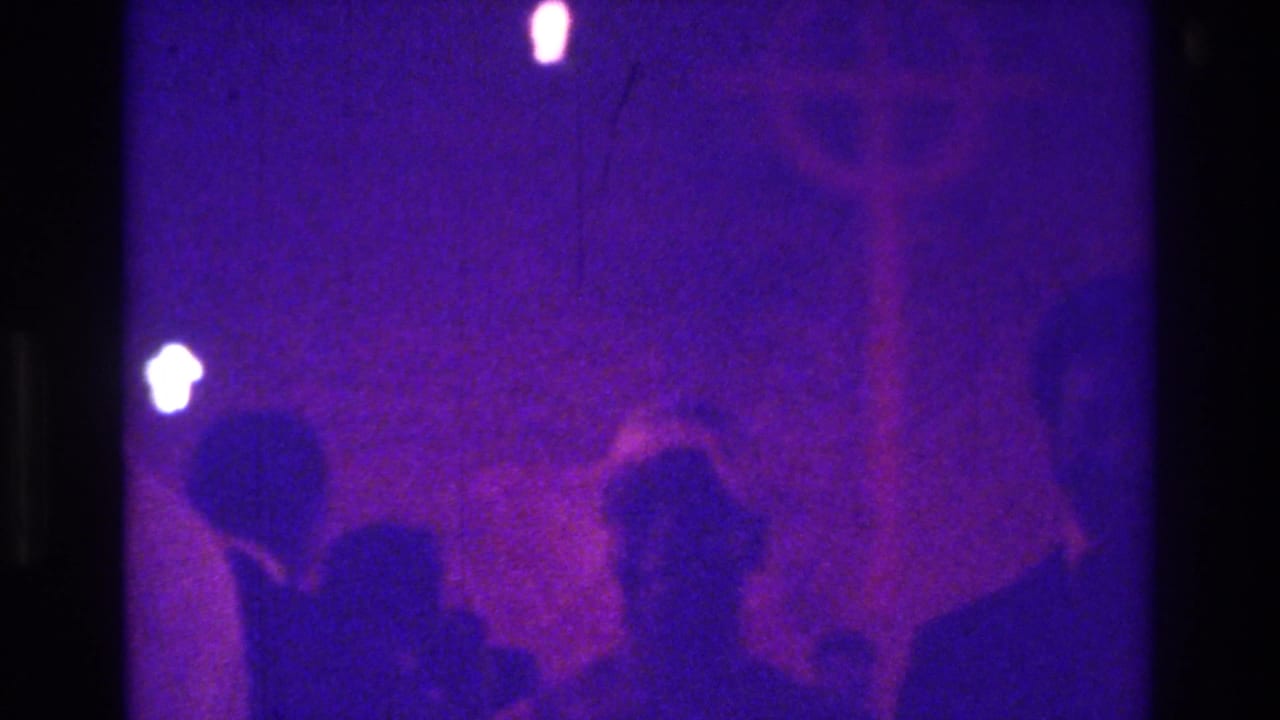 1975 Bronx home-movie frame with several silhouettes in purple light, including a person in a floppy bonnet near a cross-like form.