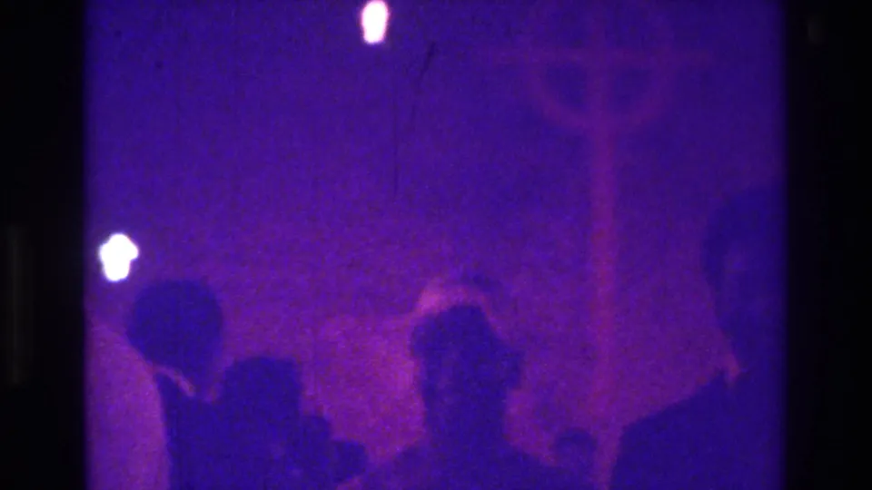 1975 Bronx home-movie frame with several silhouettes in purple light, including a person in a floppy bonnet near a cross-like form.