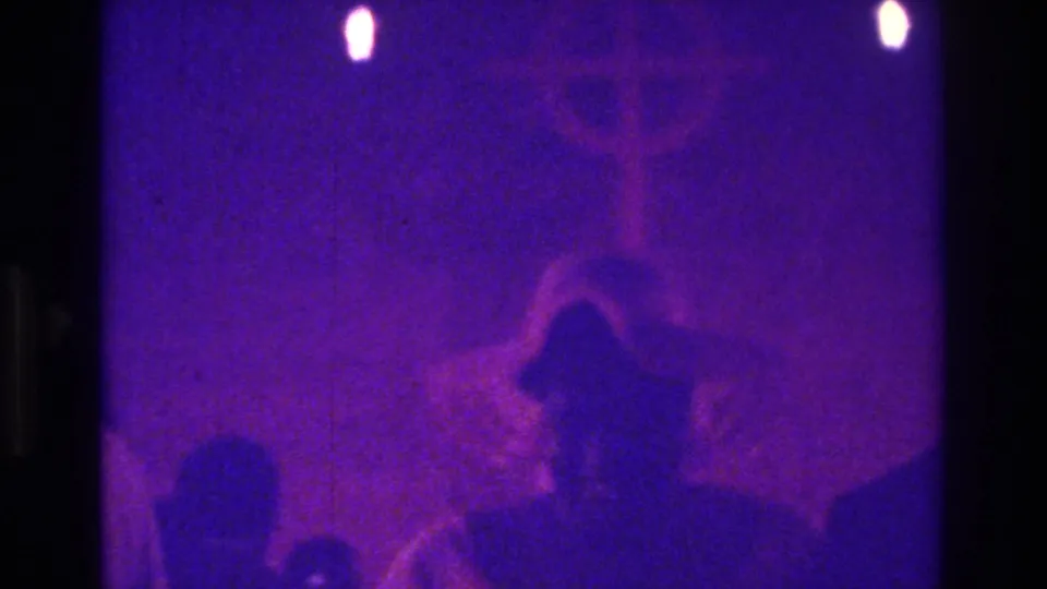 1975 Bronx home-movie frame of a bonneted silhouette in purple light, with another figure nearby and a cross-like form in the background.