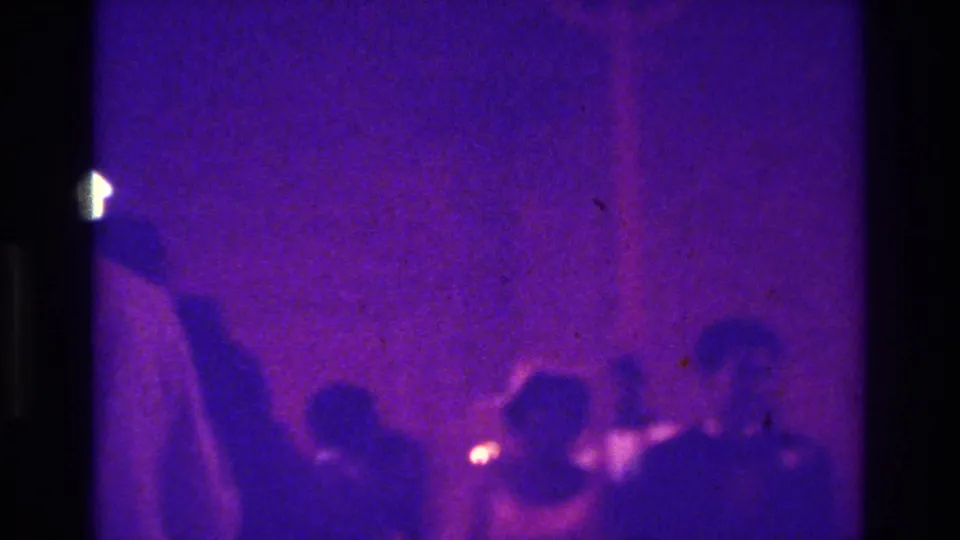 1975 Bronx church wedding frame with seated guests near the aisle, softened by purple color shift, grain, and low-light home-movie exposure.