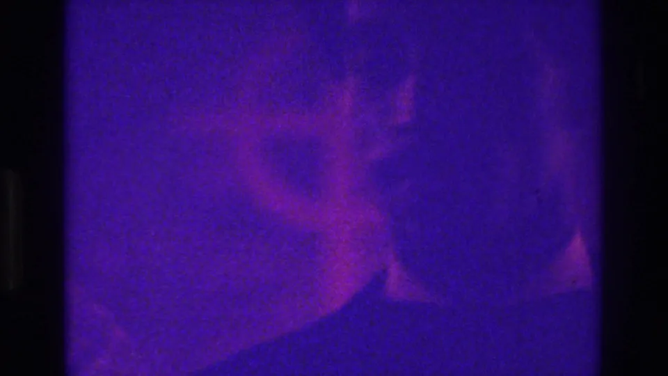 Indistinct suited person in 1975 Bronx home-movie footage, partly obscured by purple color shift, grain, and low light.