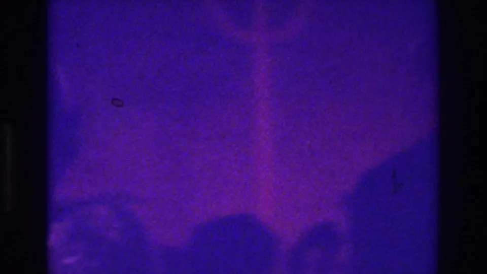 An indistinct person in formal clothing appears in purple-tinted 1975 Bronx 8mm footage, with the subject mostly lost in grain and shadow.