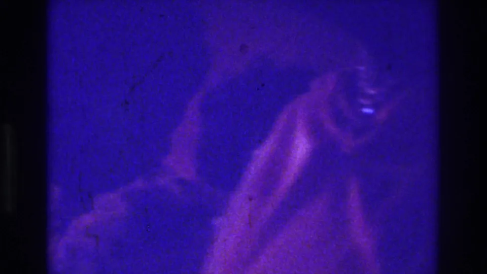 1975 Bronx 8mm frame with a shadowed figure at lower right, rendered indistinct by purple-blue light, grain, and low exposure.