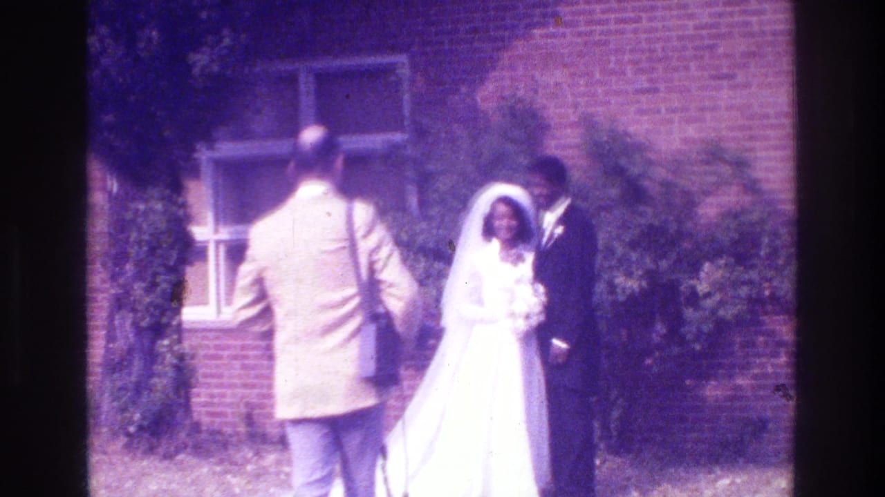 1975 Bronx home-movie frame of a bride and groom posing outdoors as a photographer in a light jacket steps into view.