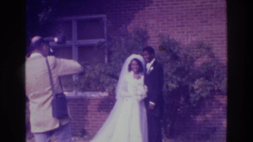 Handheld 8mm frame from the Bronx wedding scene, with the photographer partly at left and the couple posed beside the building.