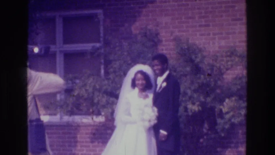 Soft 1975 Bronx wedding home-movie frame showing the bride and groom posed outdoors with the photographer partly cropped at left.
