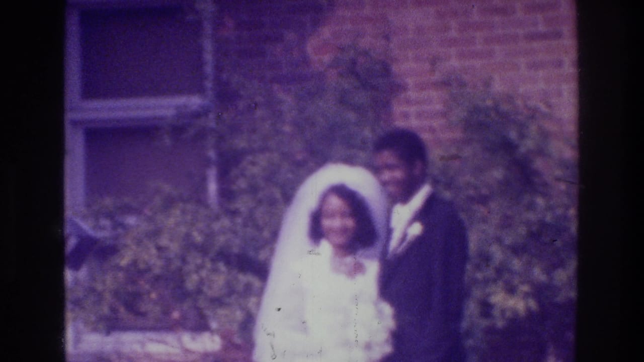 Smiling bride and groom remain posed beside shrubs and a brick wall.