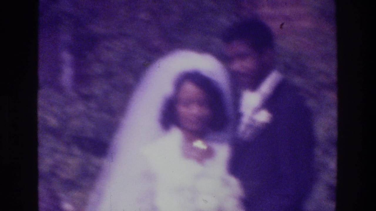 In supported 1975 Bronx home-movie footage, a bride and groom stand close together outdoors with greenery and brickwork behind them.