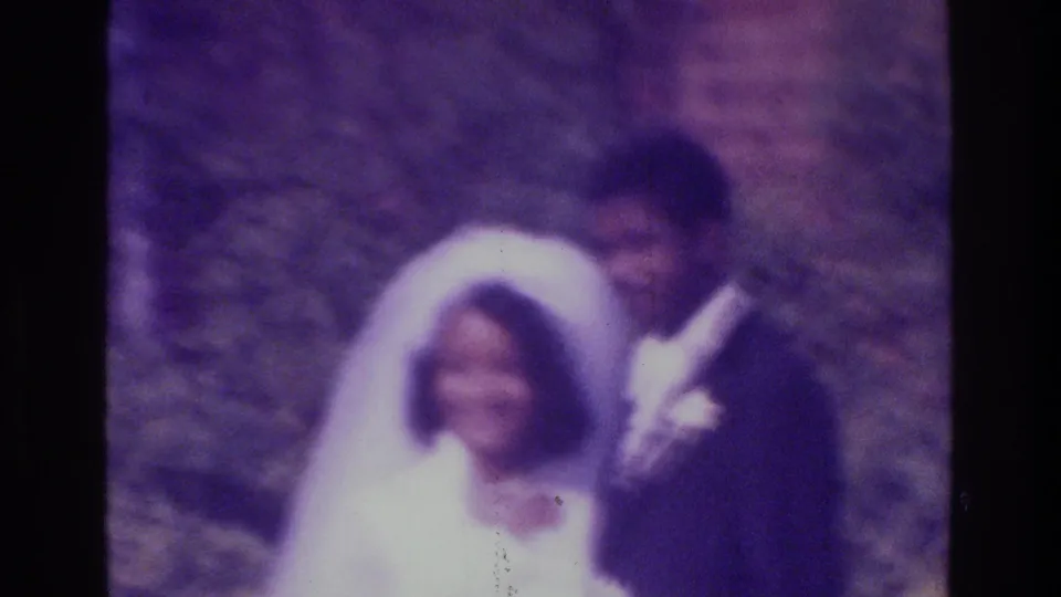 A soft-focused 1975 Bronx wedding frame shows the bride in white and the groom in a dark suit standing before foliage and brick.