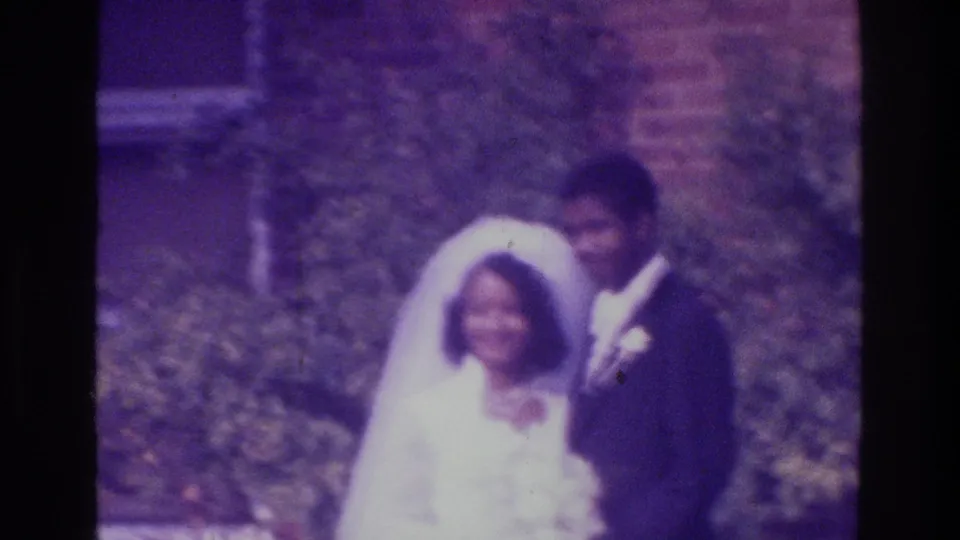 The couple smiles toward the camera in supported 1975 Bronx footage, framed by greenery and a brick exterior.