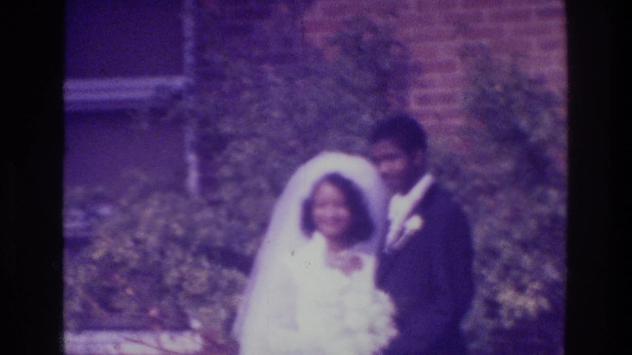 1975 Bronx home-movie frame of a bride in a white gown and veil standing with a groom in a dark suit outdoors.